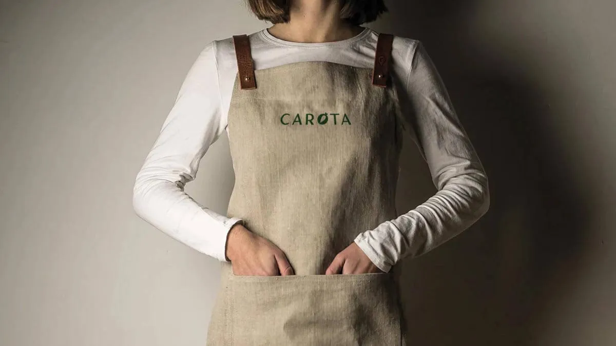 Person wearing a beige apron with brown leather straps and the word 'CAROTA' embroidered on the front pocket, hands inside the pocket.