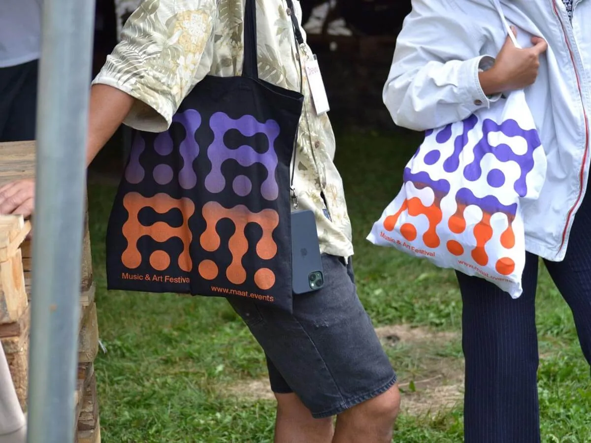 Two people holding tote bags with purple and orange abstract patterns and text promoting a Music & Art Festival.