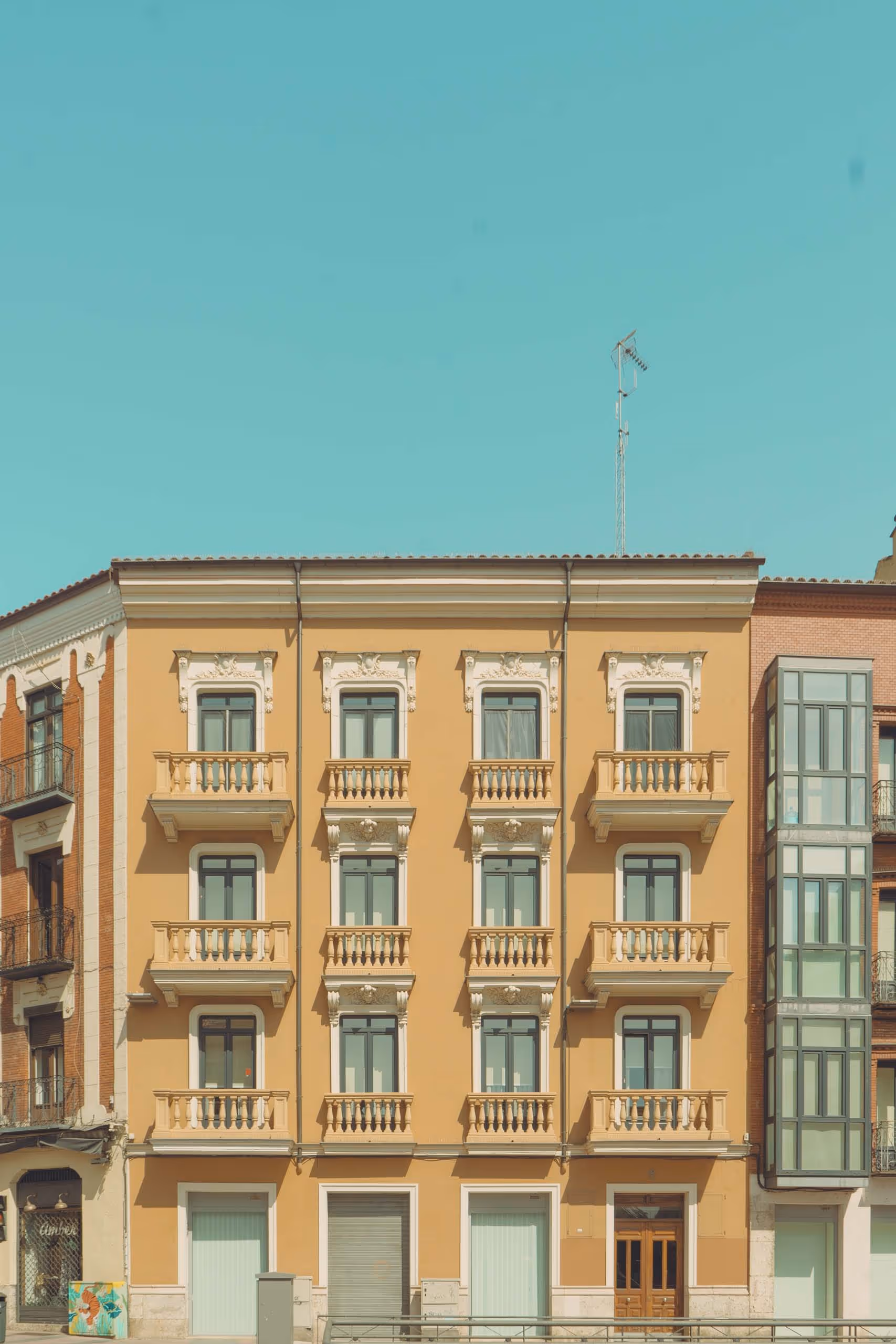 Four-story beige building with nine windows featuring decorative balconies and a wooden door under a clear blue sky.
