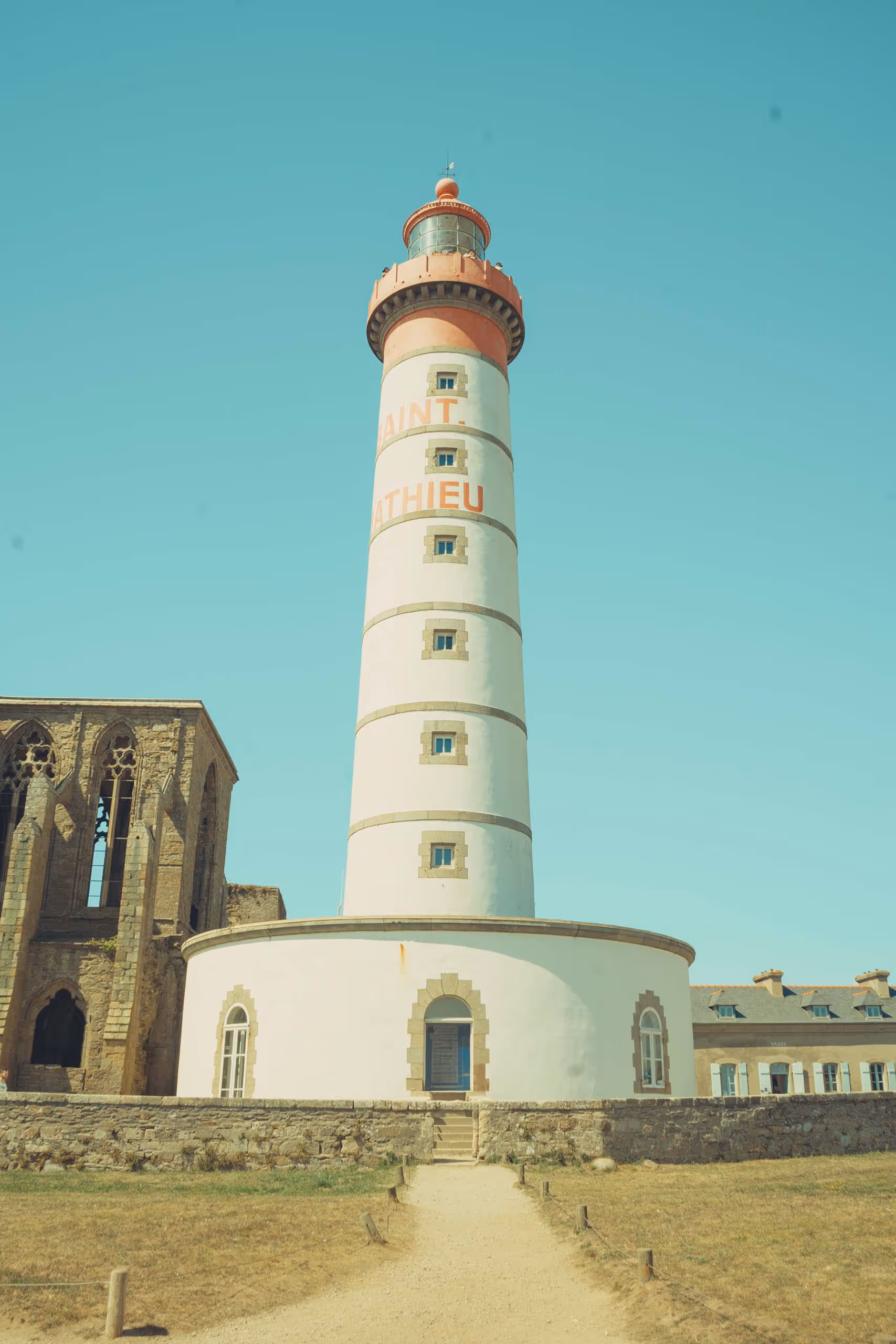 Tall white lighthouse with red accents and the text 'Saint Mathieu' against a clear blue sky, with a stone building on the left and a grassy path leading up to it.