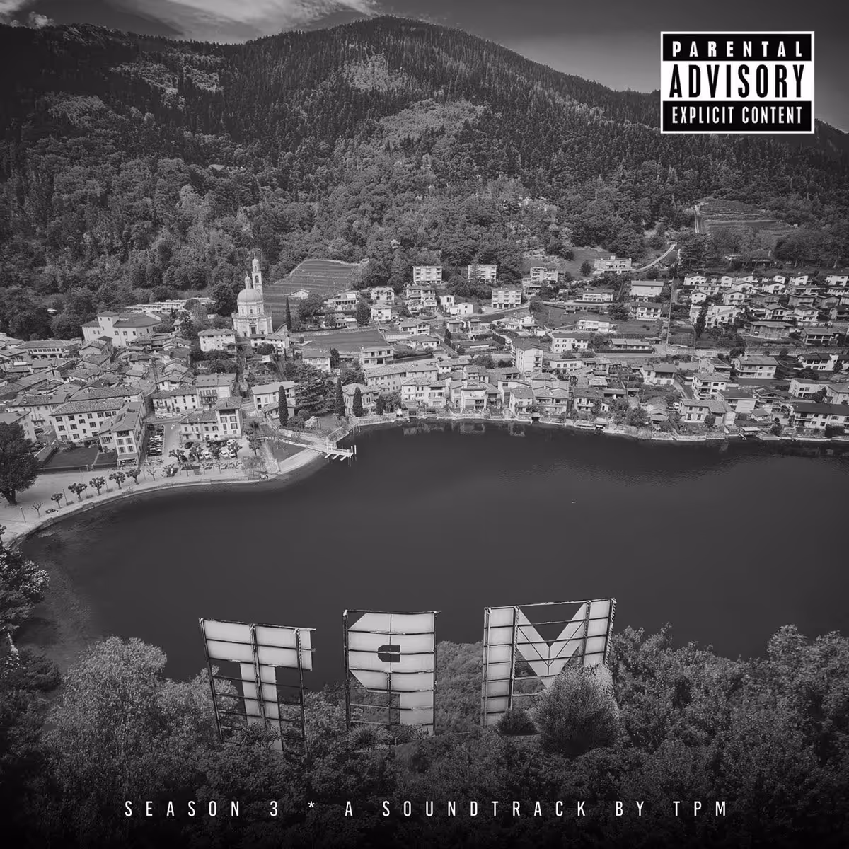 Black and white aerial view of a lakeside town with large letters TPM on a hillside and Parental Advisory label.