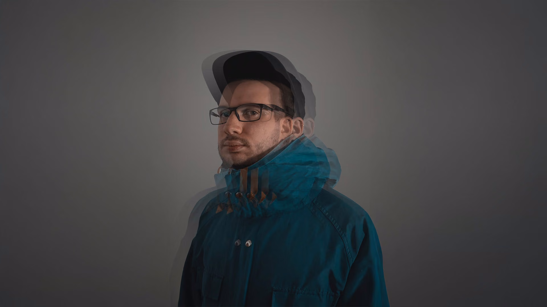Man wearing glasses, a cap, and a blue jacket with a motion blur effect showing multiple overlapping images of his head.