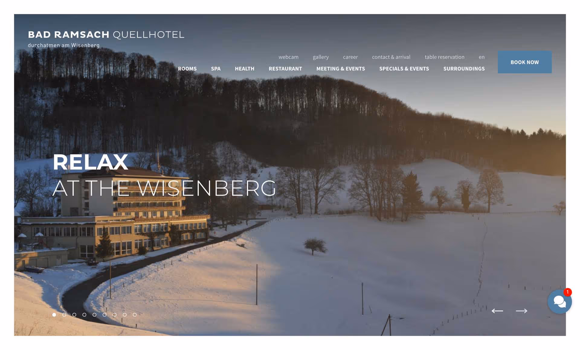 Snow-covered landscape at sunset with a large hotel building at the base of a wooded hill.