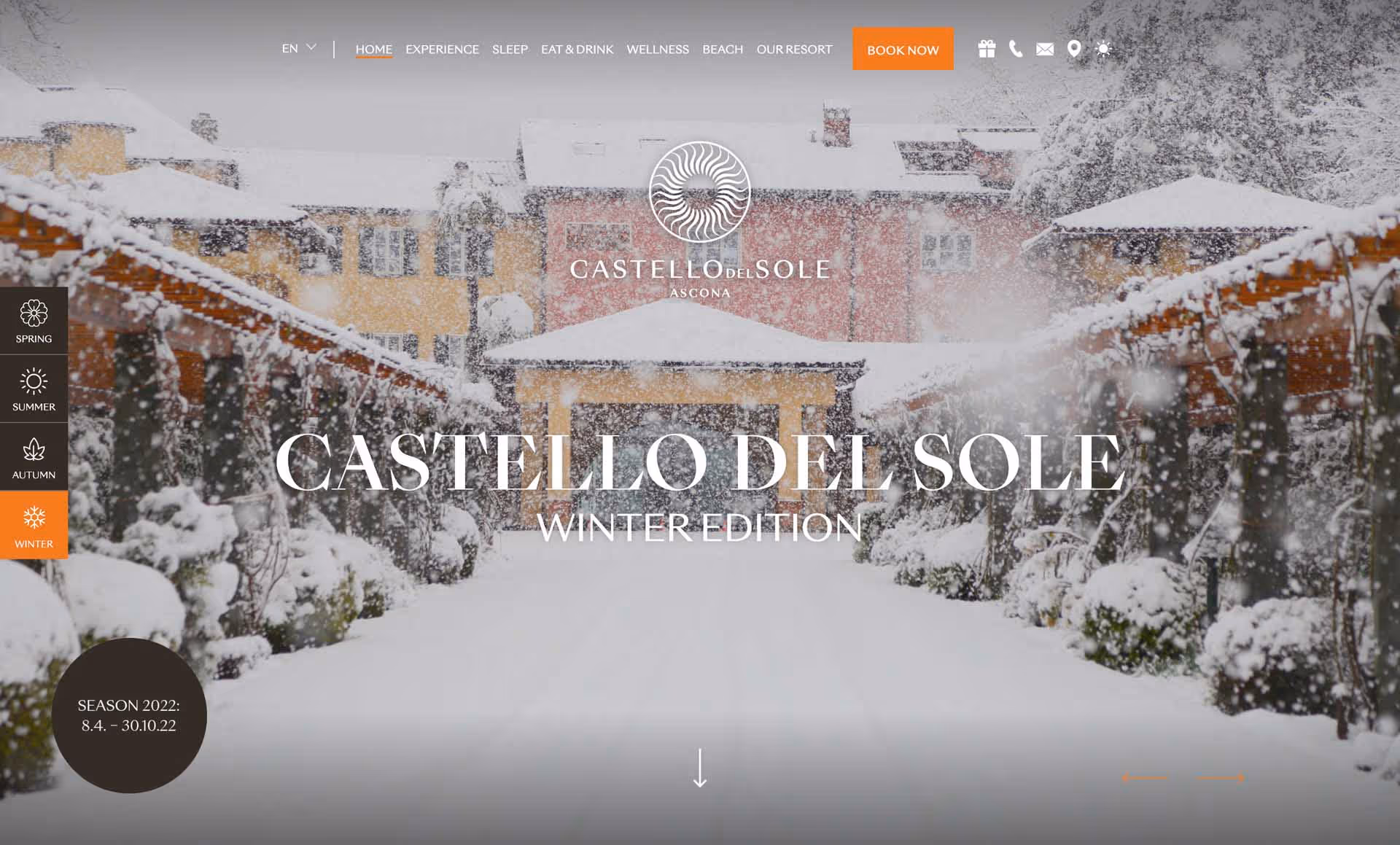 Snow-covered pathway lined with bushes and wooden pergolas leading to pastel-colored buildings during heavy snowfall at Castello del Sole resort.