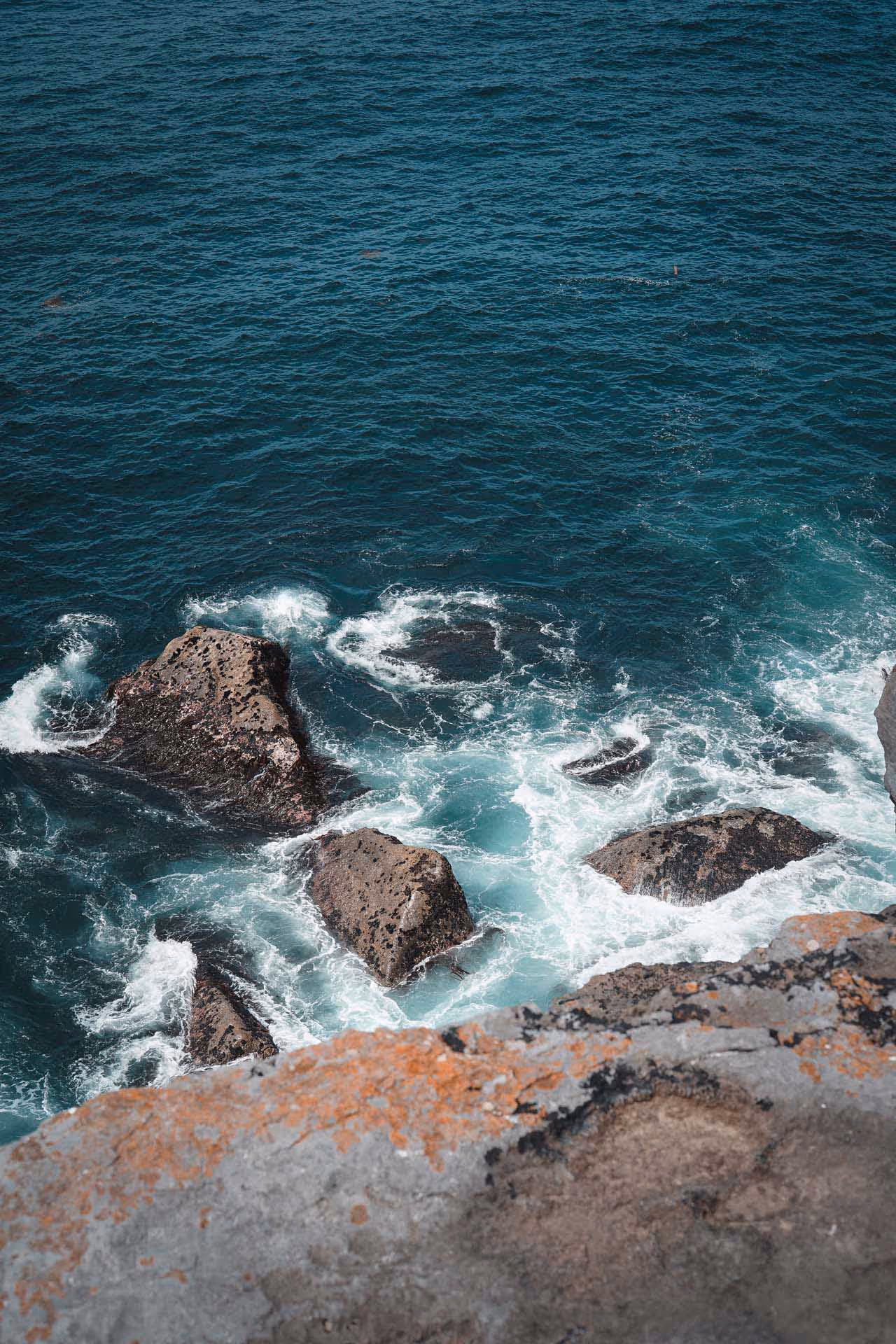 Waves crashing against large rocks along a rugged coastline with a rocky cliff edge in the foreground.