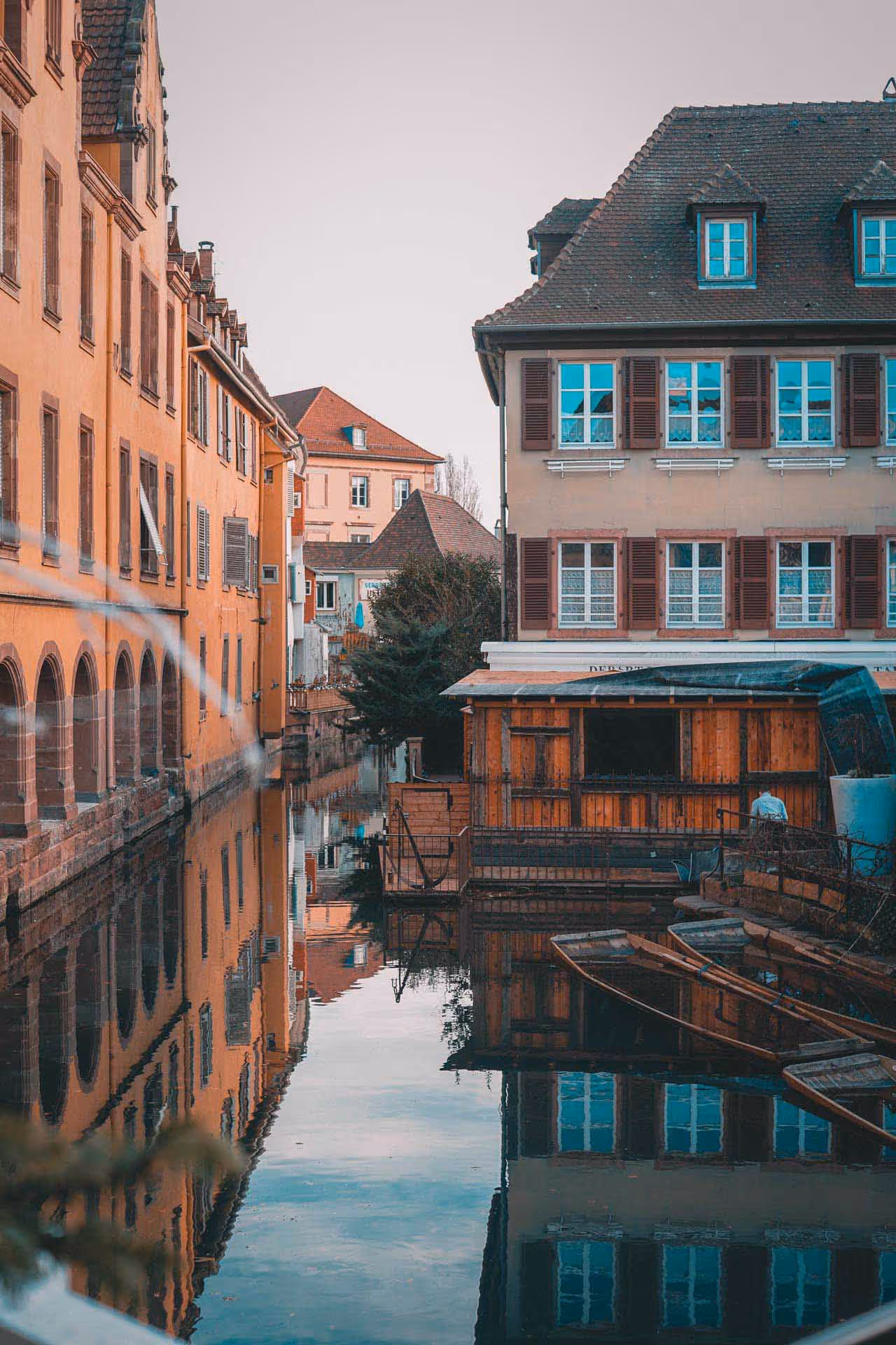 Quiet European canal with orange buildings and wooden boats reflected in calm water under a clear sky.