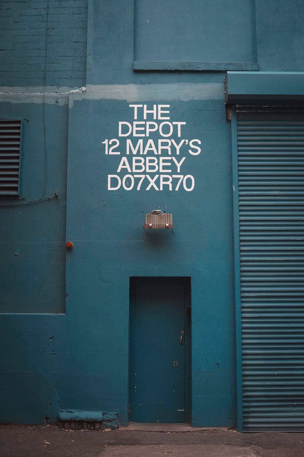 Teal-colored building wall with white text reading 'THE DEPOT 12 MARY'S ABBEY D07XR70', a closed teal door, and a large shutter door.