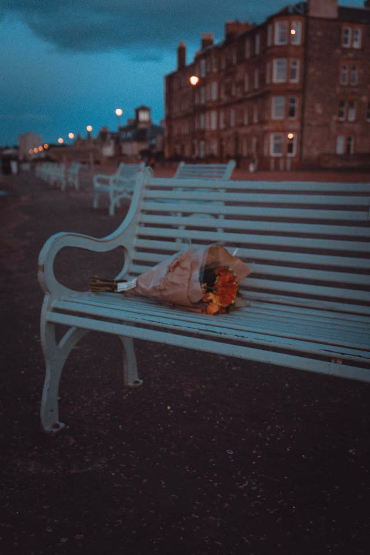 Bouquet of flowers wrapped in paper resting on a light blue bench at dusk with buildings and streetlights in the background.