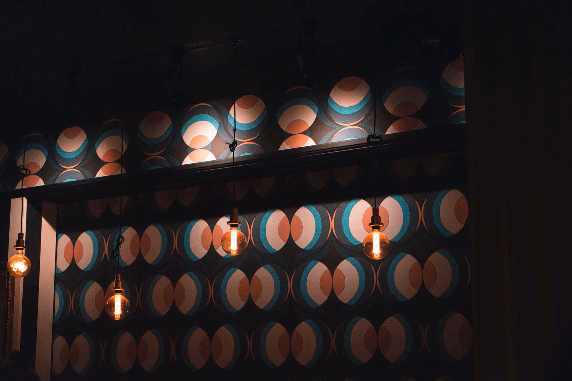 Four hanging vintage-style Edison bulbs illuminated against a dark wall with blue, brown, and white circular patterned wallpaper.