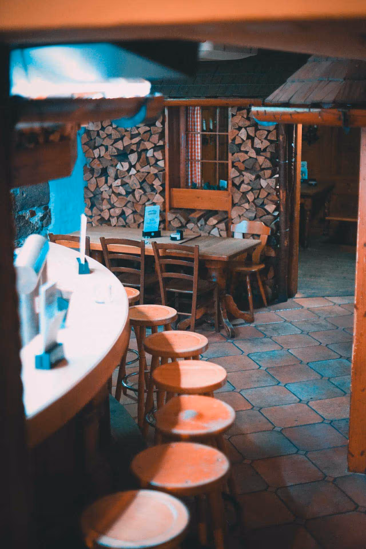 Cozy wooden interior of a rustic bar with round wooden stools lined up along a curved counter and stacked firewood against the wall.