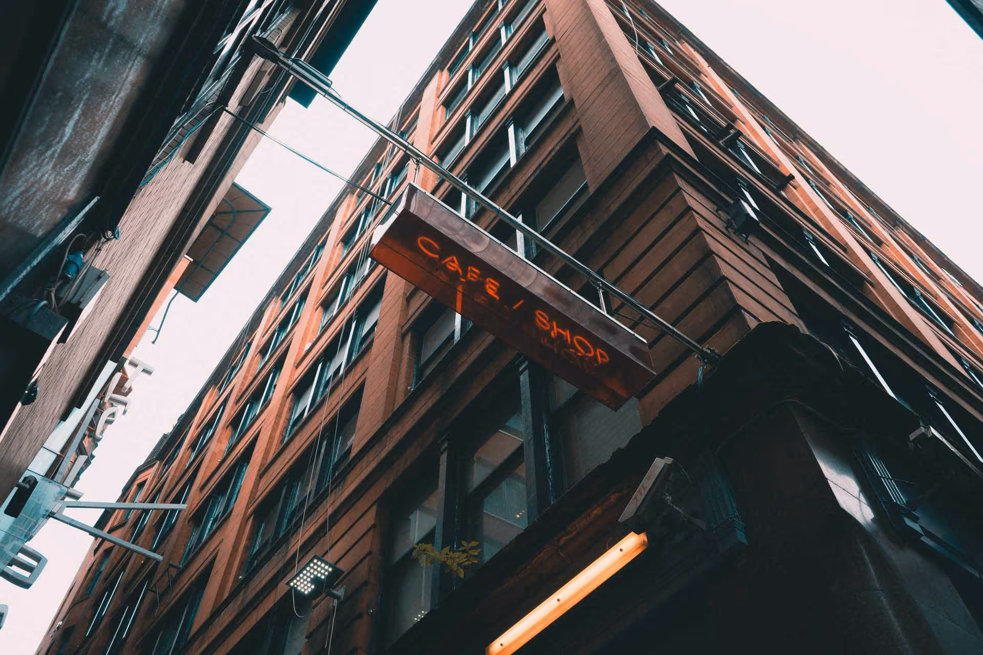 Upward view of a brick building corner with a hanging sign reading 'Cafe / Shop' in red neon lights.