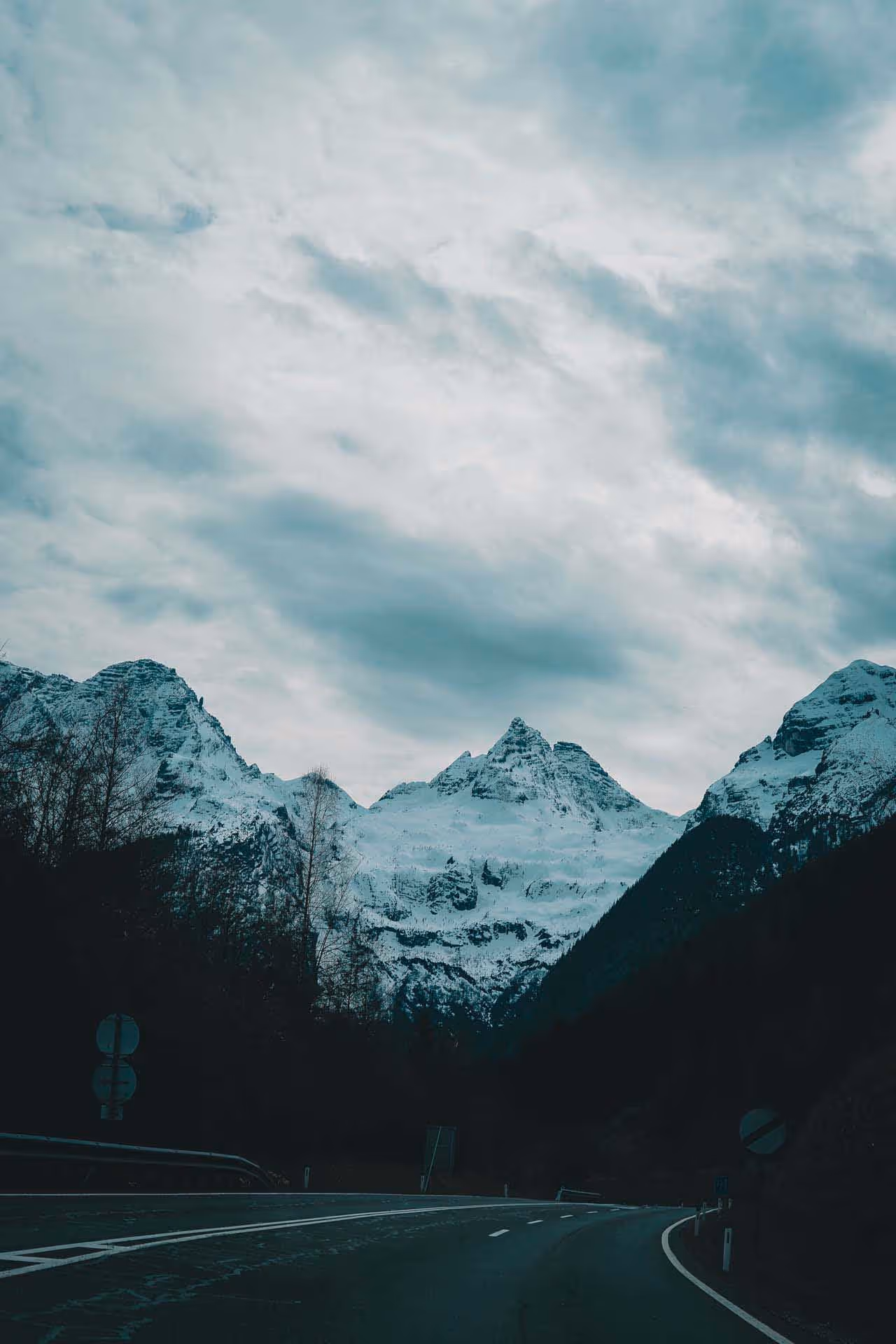 Curved road leading through a dark valley toward snow-covered mountain peaks under a cloudy sky.