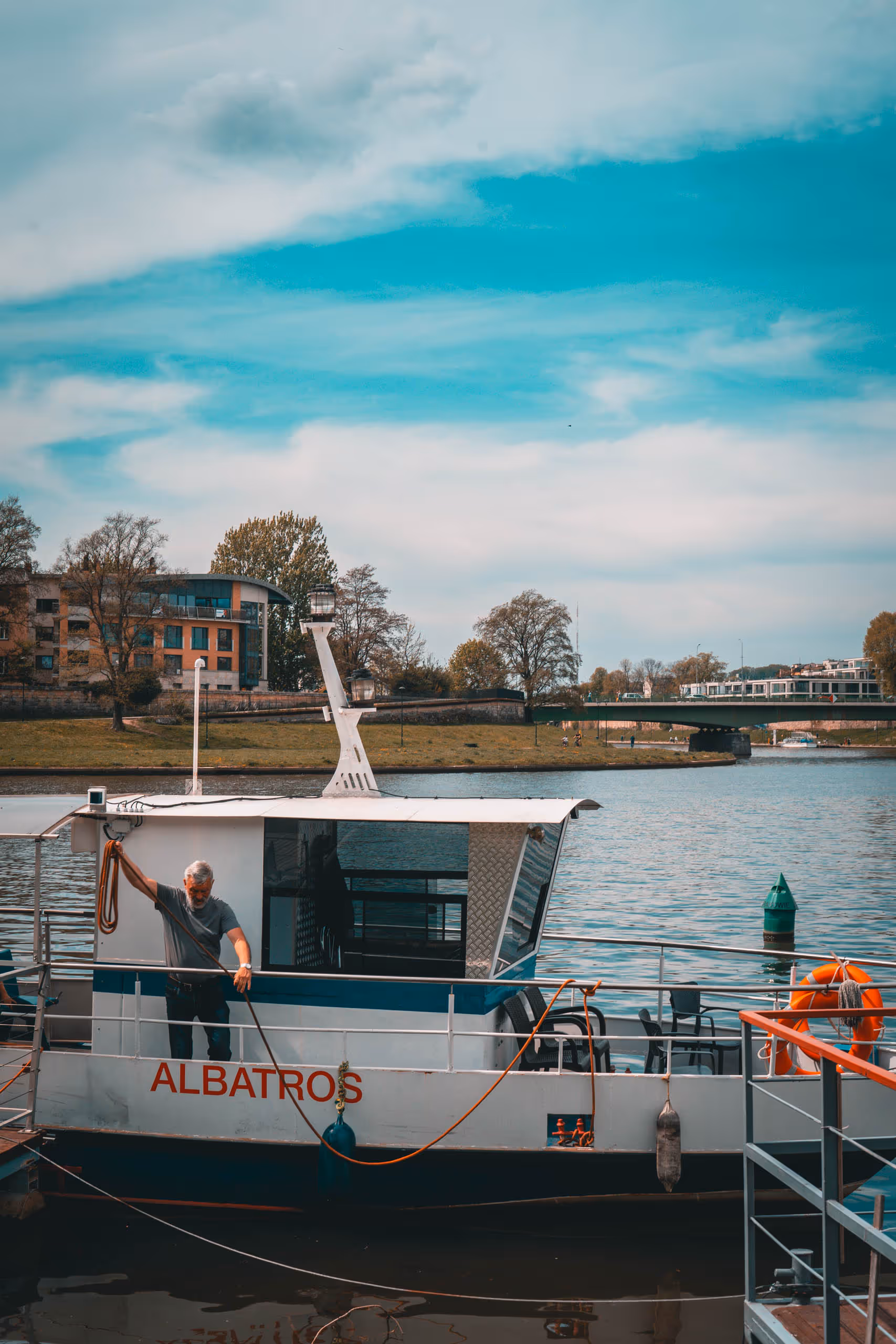 Man with gray hair and beard handling a rope on the deck of a white boat named Albatros docked on a river with trees and buildings in the background.
