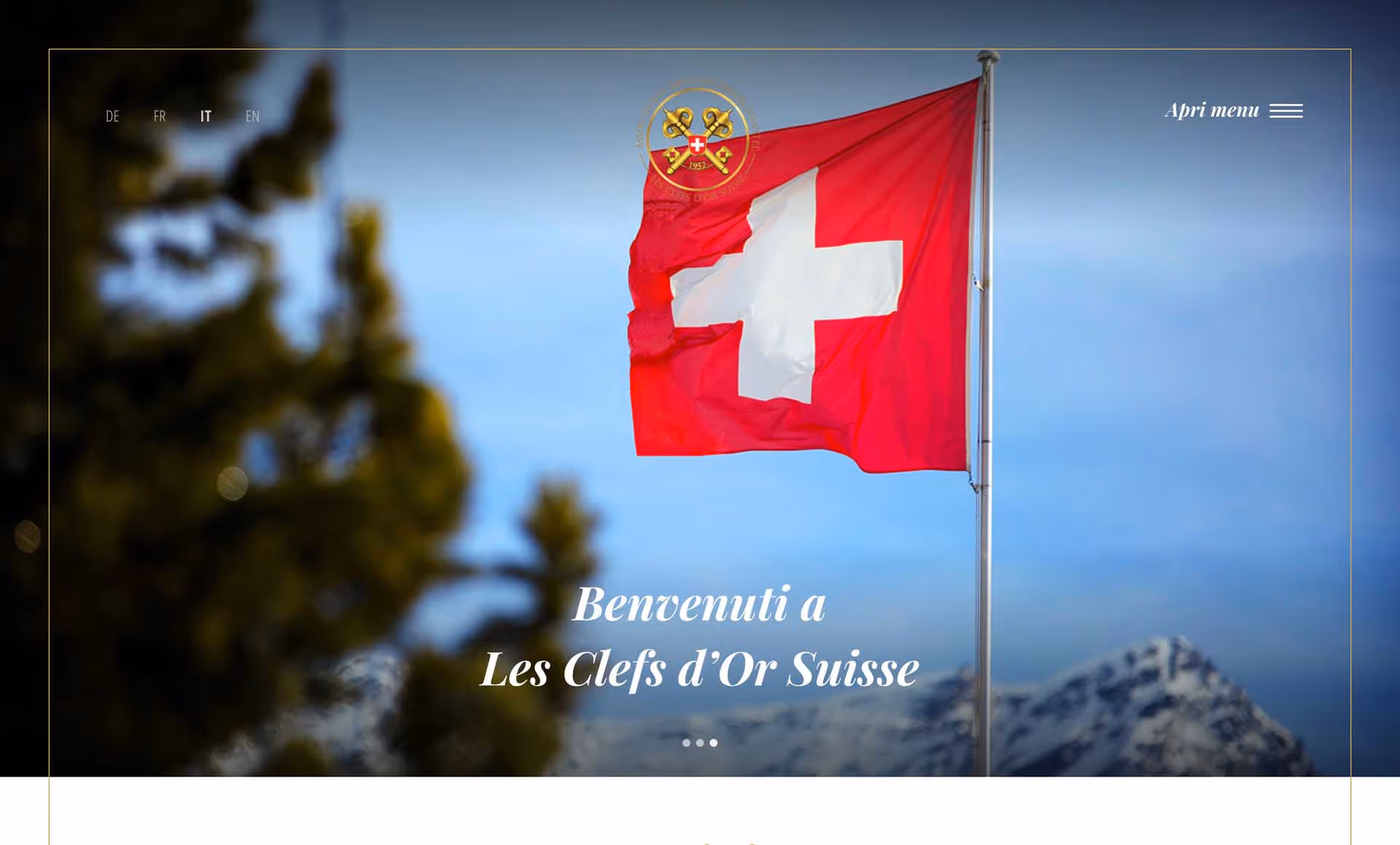 Swiss flag waving on a flagpole with snowy mountains in the background and trees blurred in the foreground.