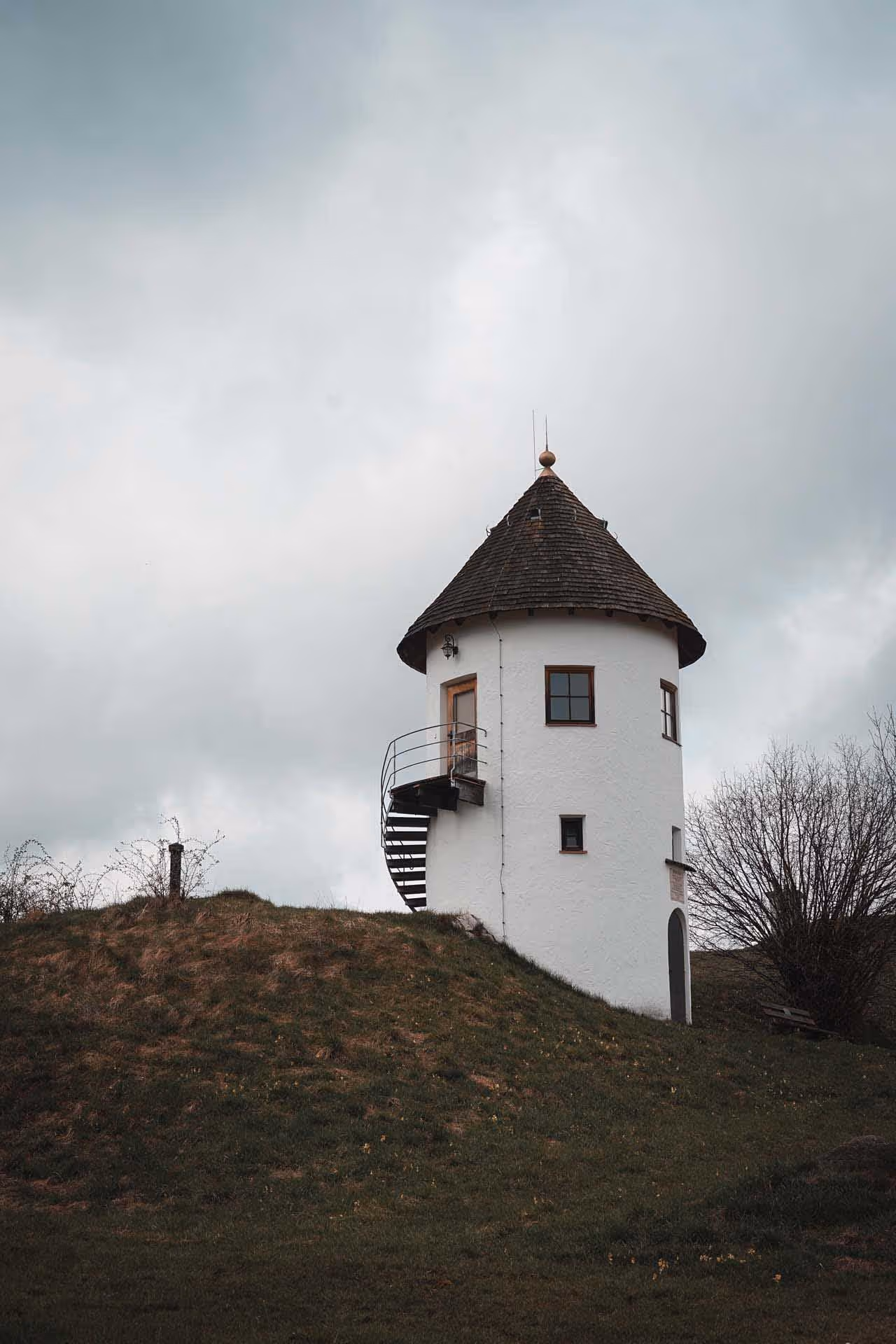 Small white cylindrical tower with a conical wooden roof and an external spiral staircase on a grassy hill under a cloudy sky.