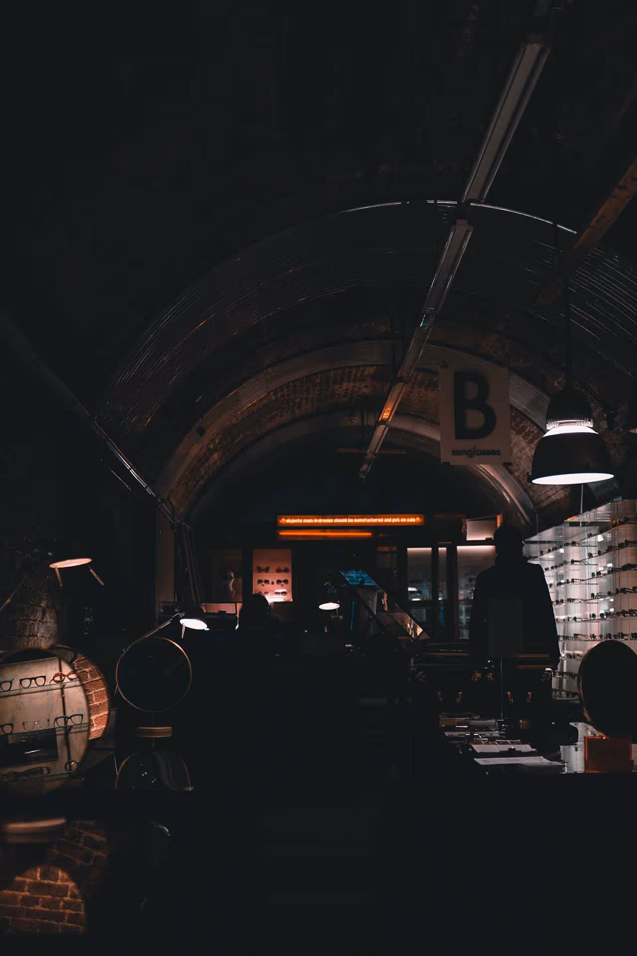 Dimly lit underground store with arched brick ceiling displaying sunglasses and eyewear under pendant and desk lamps.