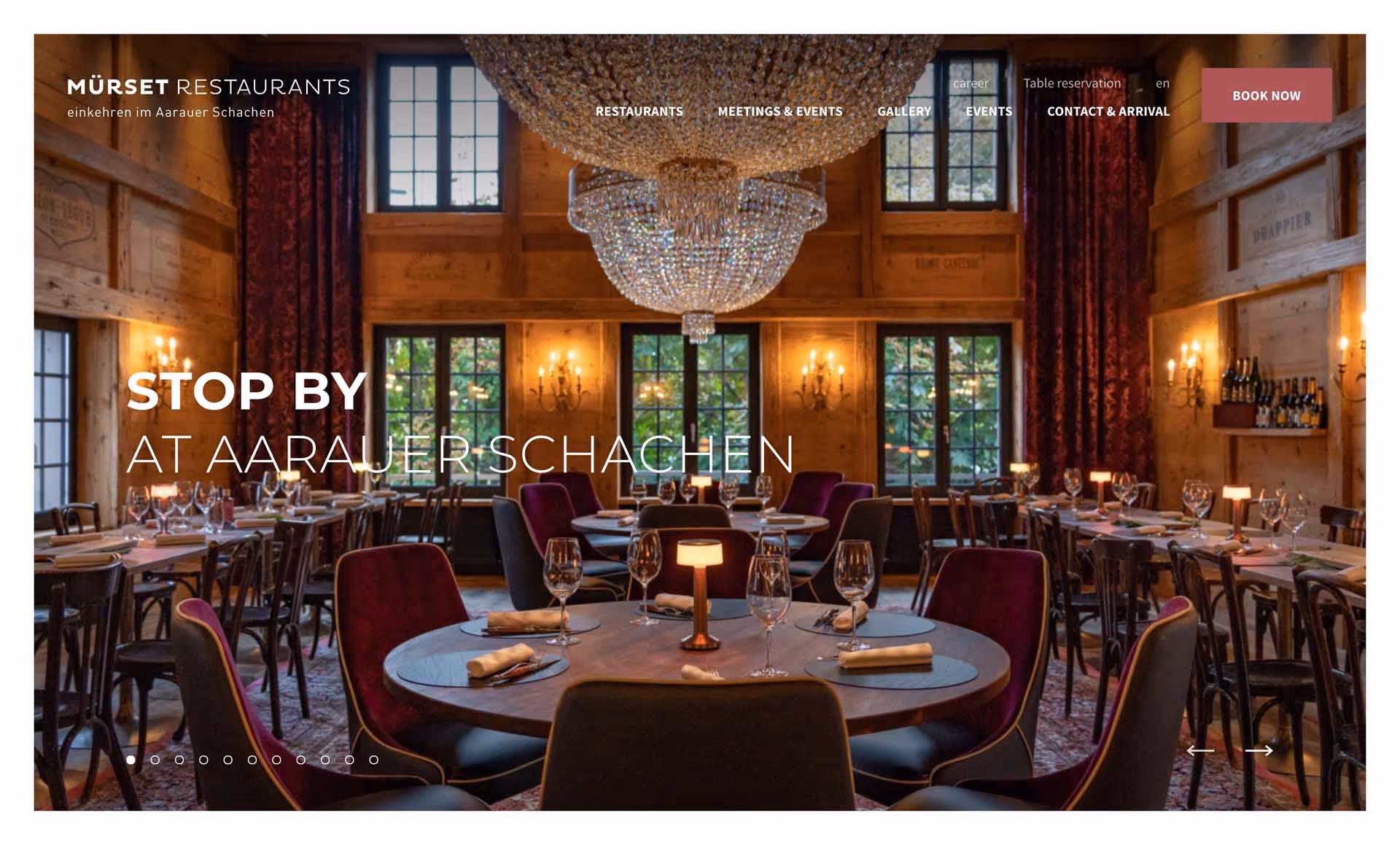 Elegant restaurant interior with a large chandelier, wooden walls, round and rectangular tables set with wine glasses and napkins.