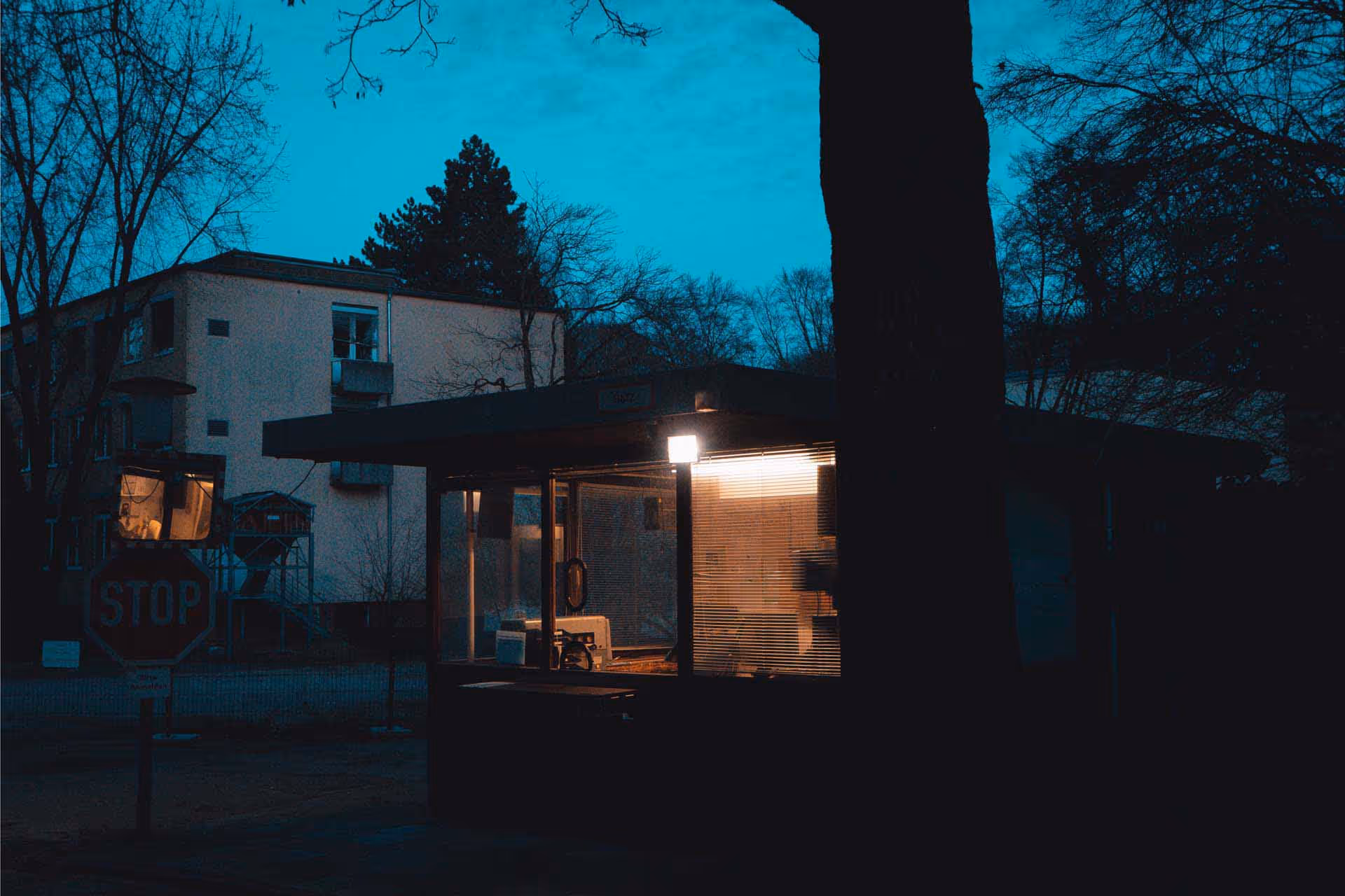 Small illuminated security or guard booth at dusk with a stop sign nearby and bare trees around.