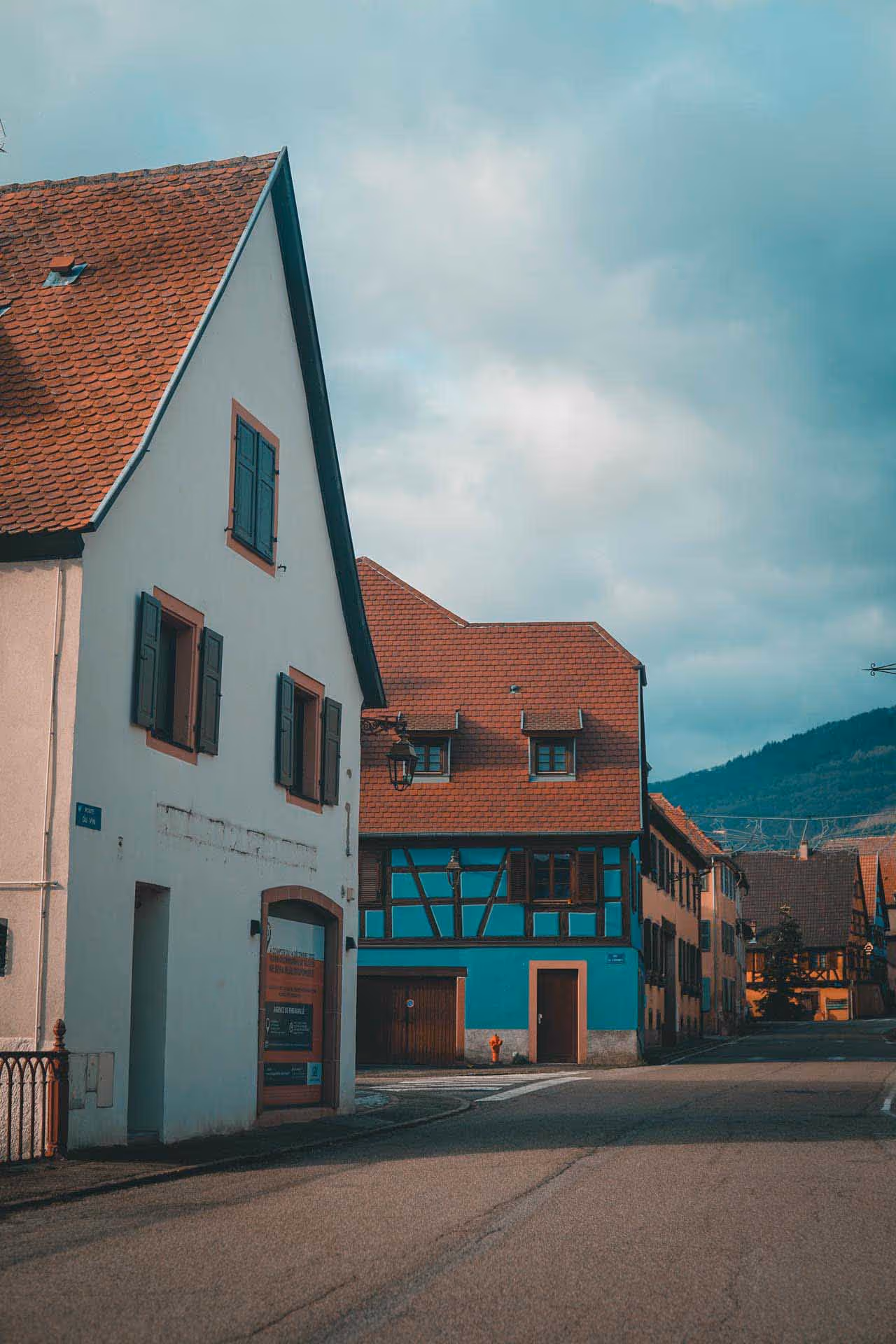 Quiet street with traditional European houses featuring red-tiled roofs and wooden shutters under a cloudy sky.