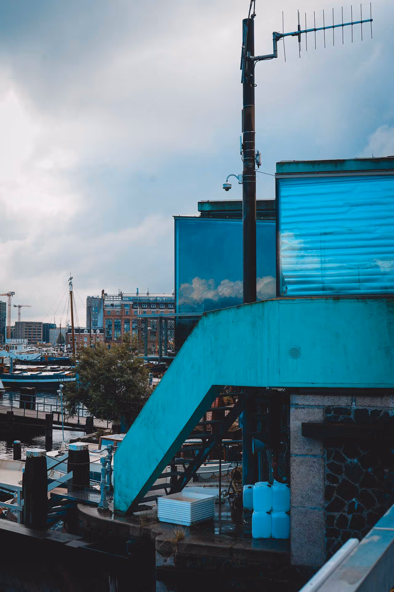 Harbor scene with blue industrial stair structure, boats docked, and urban buildings under cloudy sky.