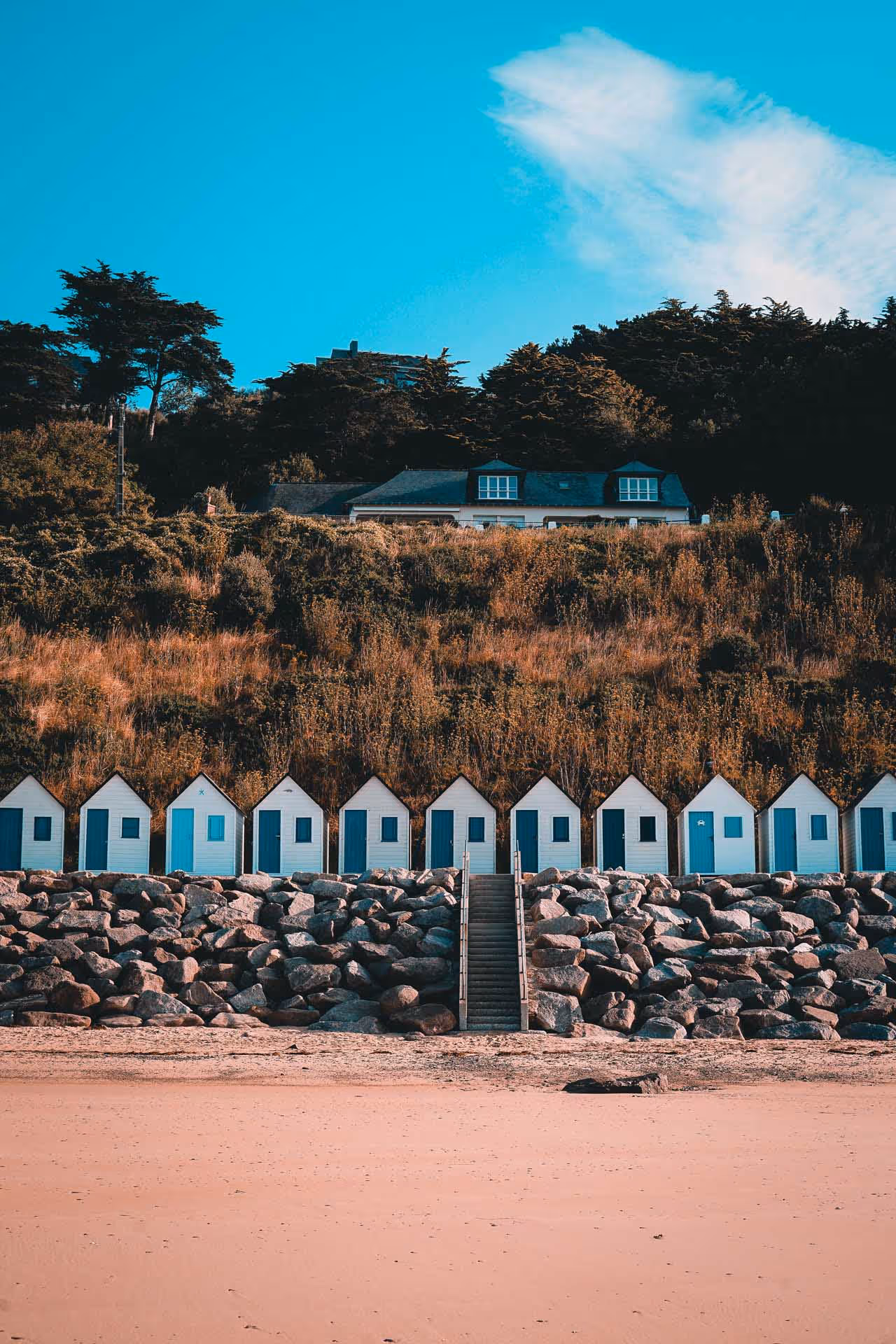 Row of small white beach huts with blue doors set on rocks below a grassy hillside and trees under a clear blue sky.
