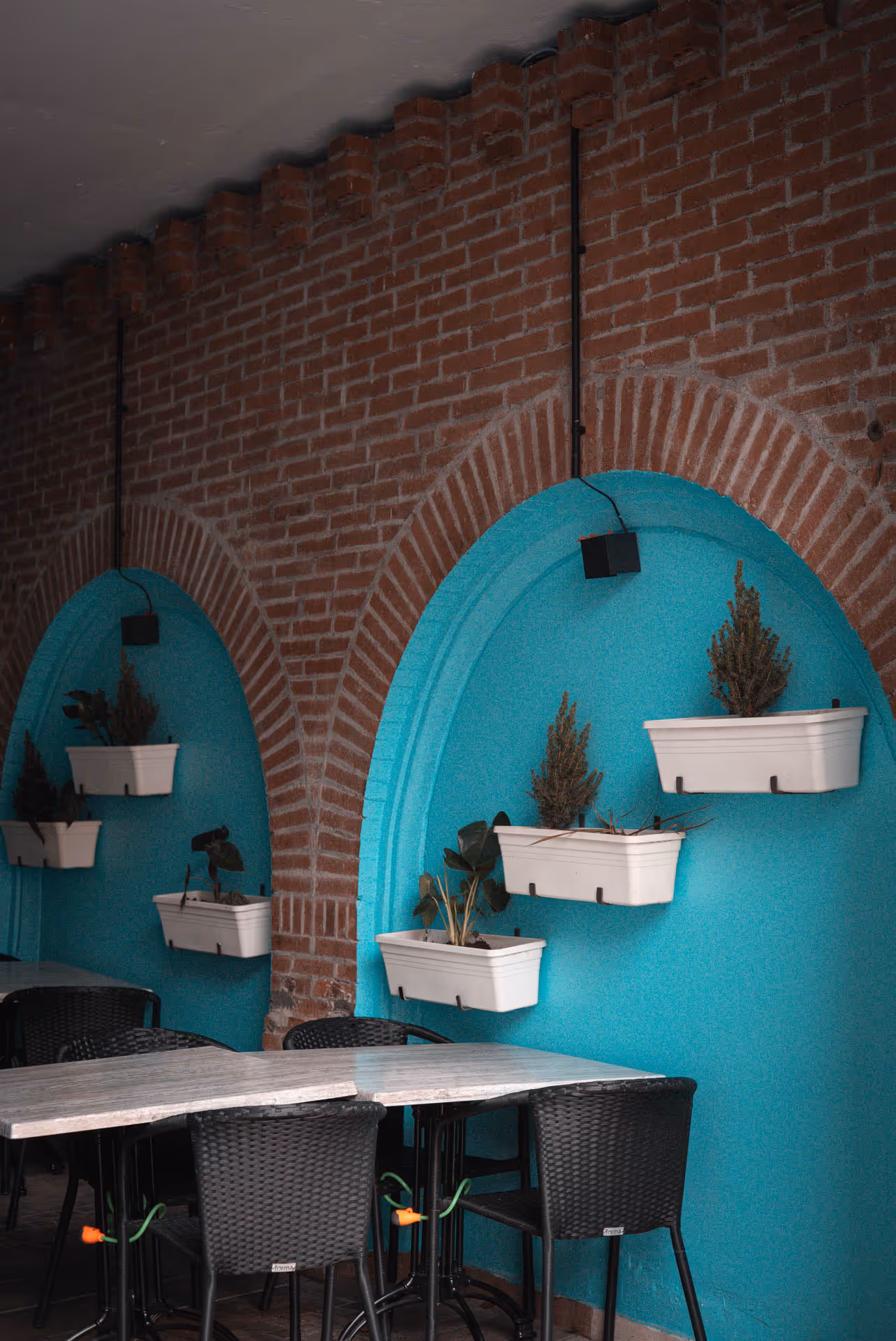 Indoor seating area with black wicker chairs and tables under brick arches painted turquoise with white wall planters containing small plants.