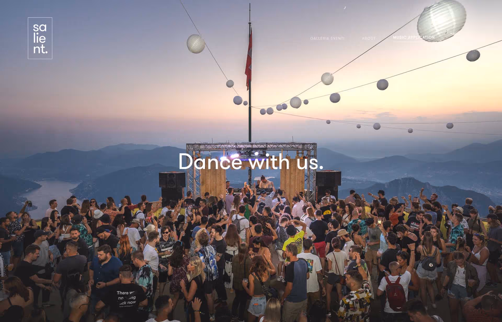 Large crowd dancing at an outdoor DJ event with mountainous landscape and sunset in the background.