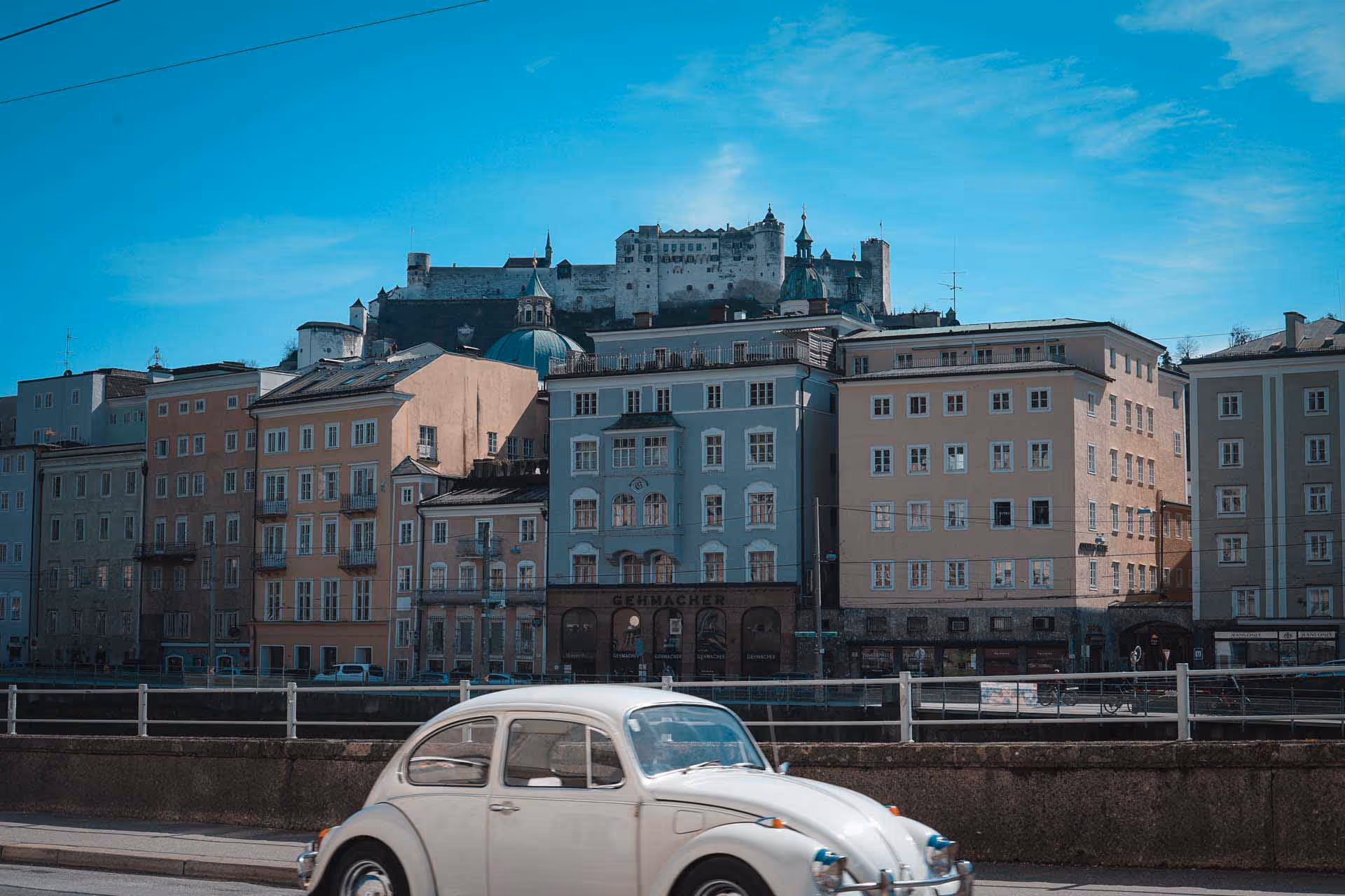 White vintage Volkswagen Beetle driving past colorful buildings with a castle on a hill in the background under a blue sky.