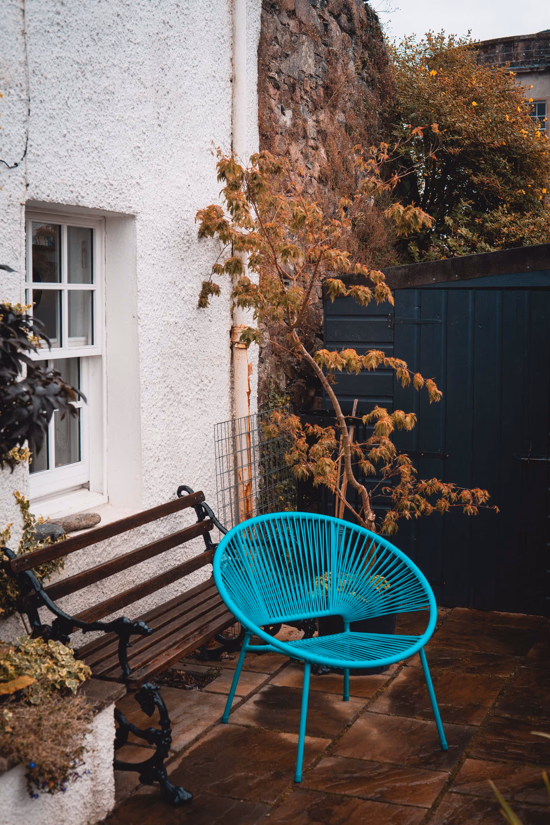 Outdoor patio with a turquoise round string chair, a wooden bench, and a small tree against white and stone walls.