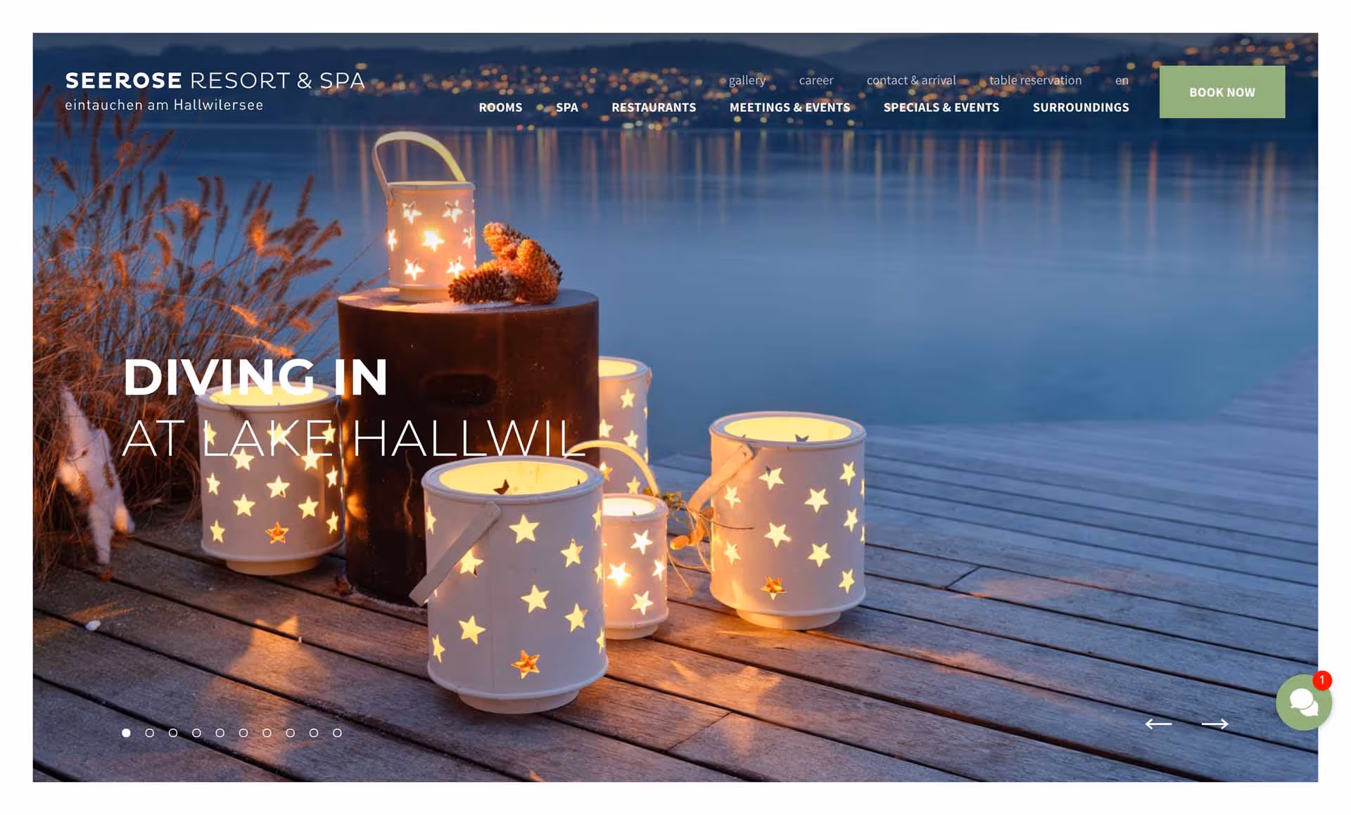 Lit white lanterns with star cutouts placed on a wooden dock by a calm lake during twilight.