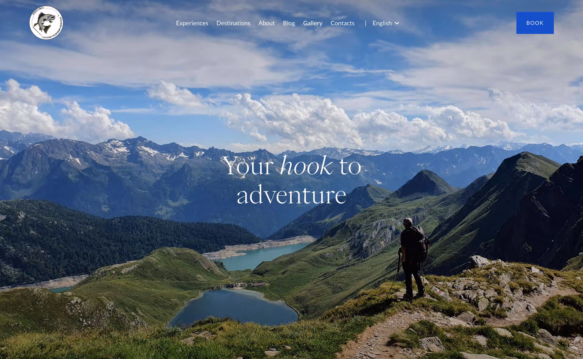 Hiker with backpack standing on a mountain trail overlooking lakes and rugged mountain peaks under a blue sky with clouds.