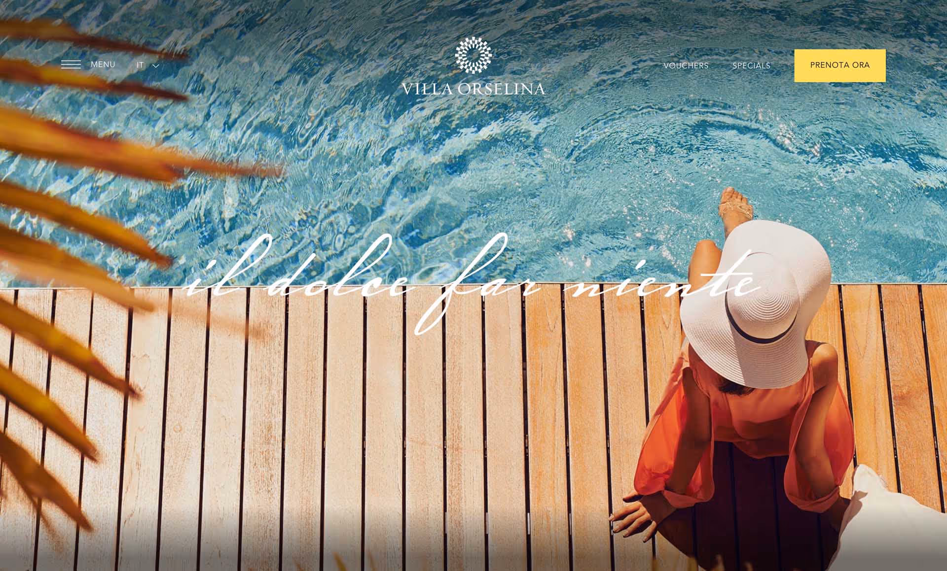 Woman in a white sunhat and orange dress sitting on a wooden deck by a clear swimming pool.