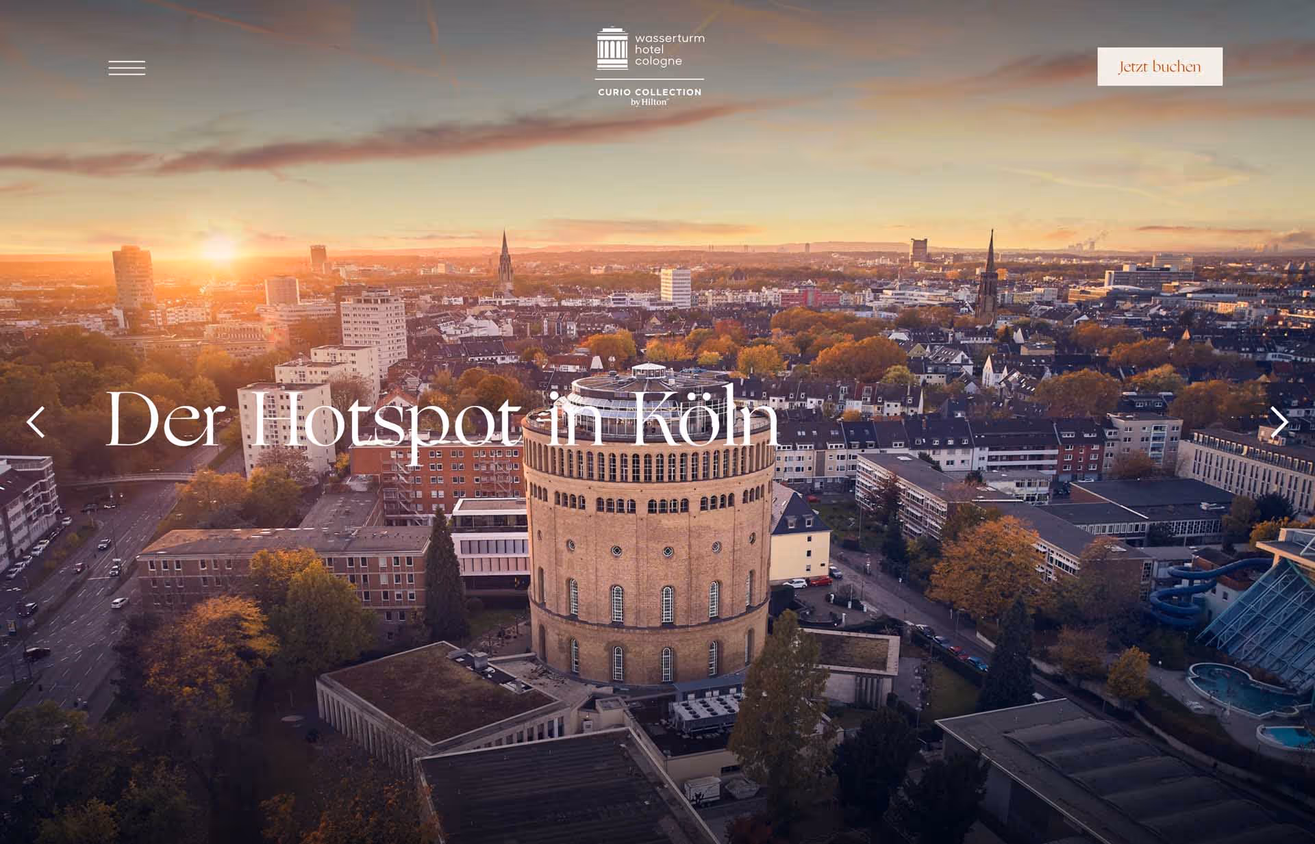 Aerial view of Cologne city at sunset featuring the historic round Wasserturm water tower surrounded by buildings and autumn trees.